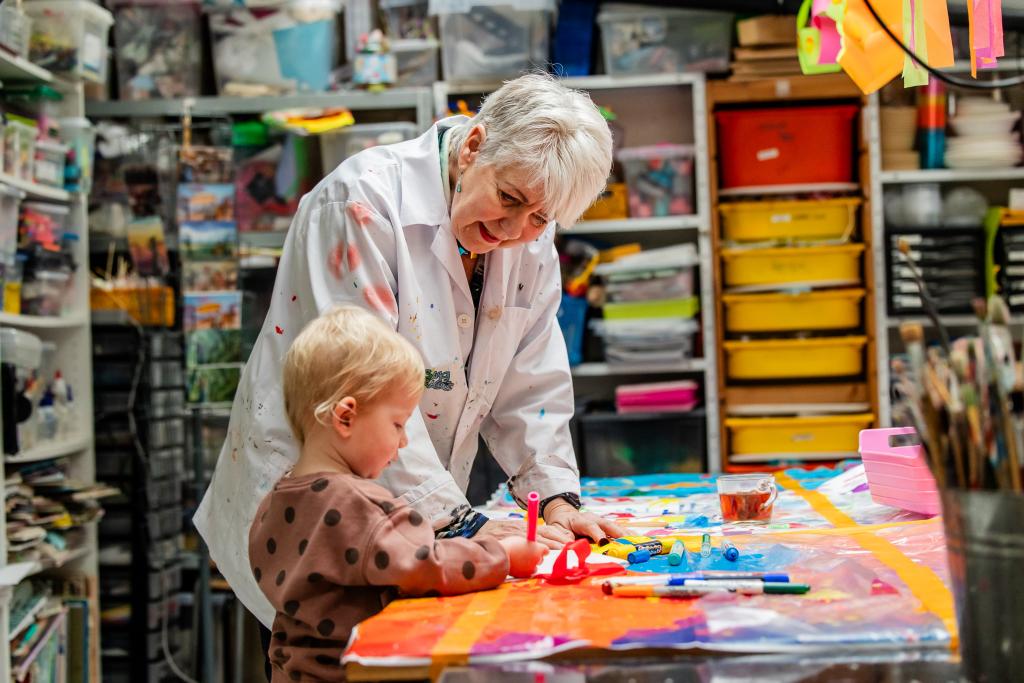 Een vrouw en jong kind zijn bezig aan een tafel in een atelier. Het kind heeft een stift vast. De vrouw kijkt naar wat het kind doet.