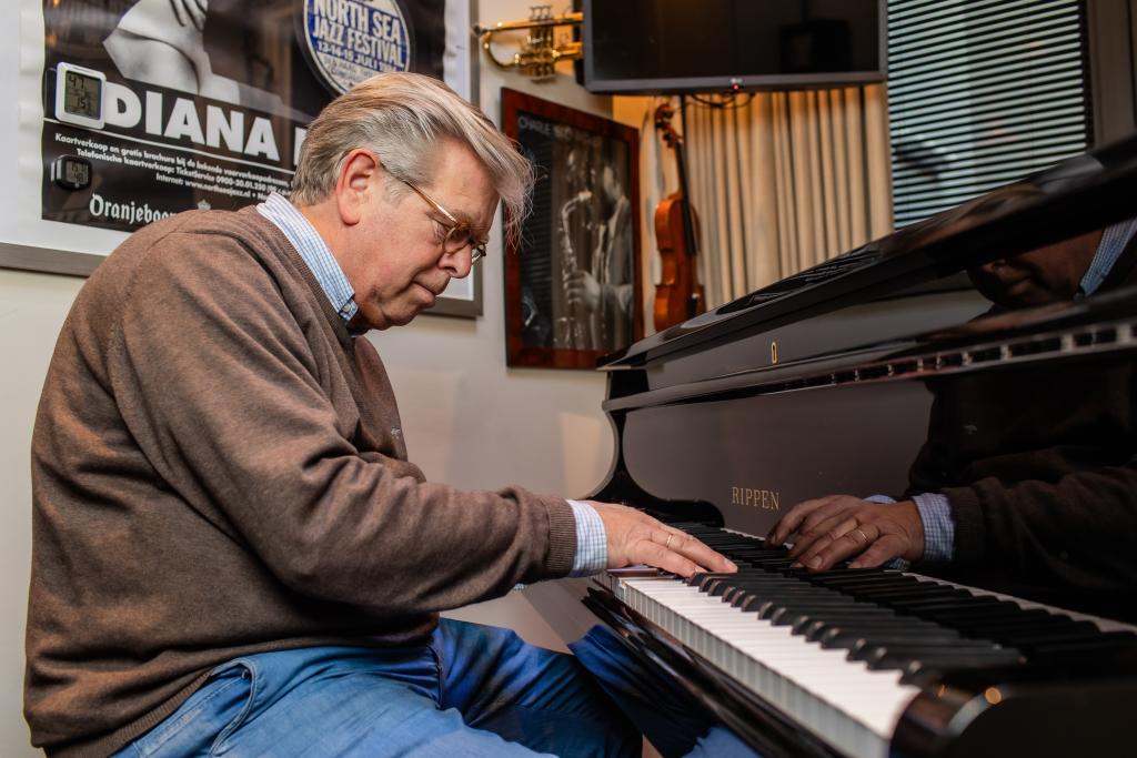 Een man zit achter een piano. Hij speelt erop. Op de achtergrond hangt een muziekposter en een viool aan de muur.