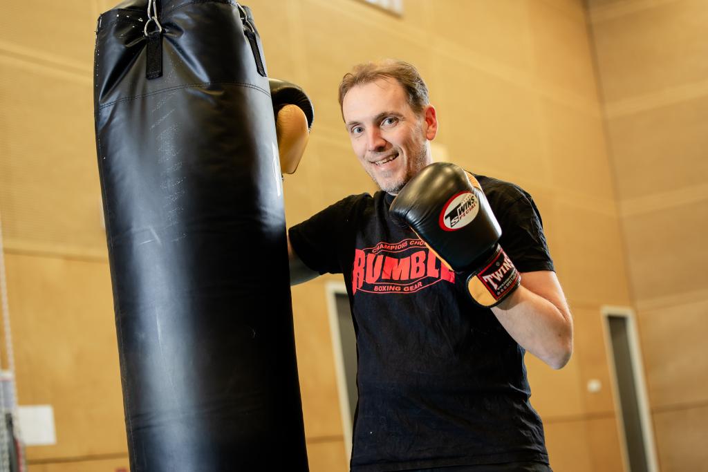 Een man staat in een sporthal. Hij heeft bokshandschoenen aan en staat bij een bokszak.